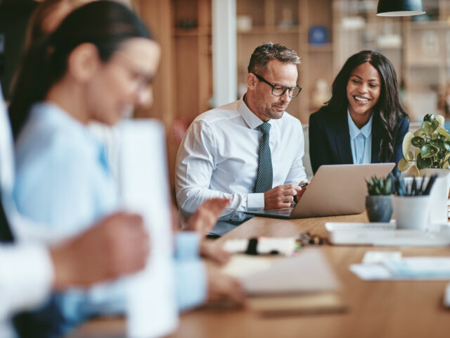 Two smiling diverse businesspeople using a laptop together at wo Two diverse businesspeople smiling while working on a laptop together at the end of a boardroom table in an office.